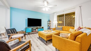 Ein Wohnzimmer mit einer Couch, Hocker und Sesseln, einem Flachbildfernseher und einem Deckenventilator.