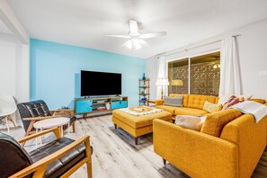 Ein Wohnzimmer mit einer Couch, Hocker und Sesseln, einem Flachbildfernseher und einem Deckenventilator.
