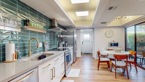 Uma cozinha moderna com armários brancos, fogão de aço inoxidável e painel de parede verde. Há uma área de jantar com mesa redonda e cadeiras de madeira.