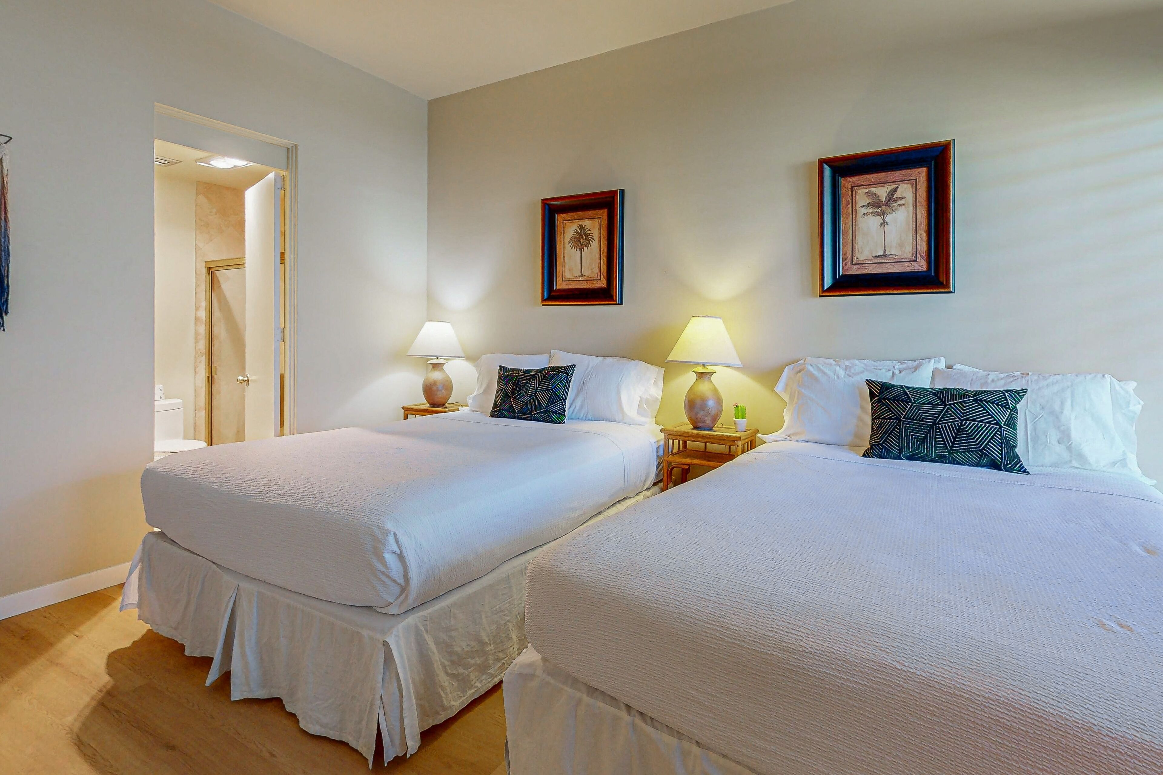 Une chambre d’hôtel avec deux lits, chacun recouvert d’une couvre-lit blanche et agrémenté d’oreillers assortis. On y trouve deux tables de chevet avec des lampes, et les murs sont décorés d’œuvres d’art encadrées.