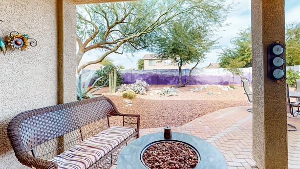 A patio with a wicker sofa, a round table with a fire pit, and a view of a landscaped garden. - Desert retreat with enclosed yard, instruments, alfresco dining, grill, & W/D (San Tan Valley)