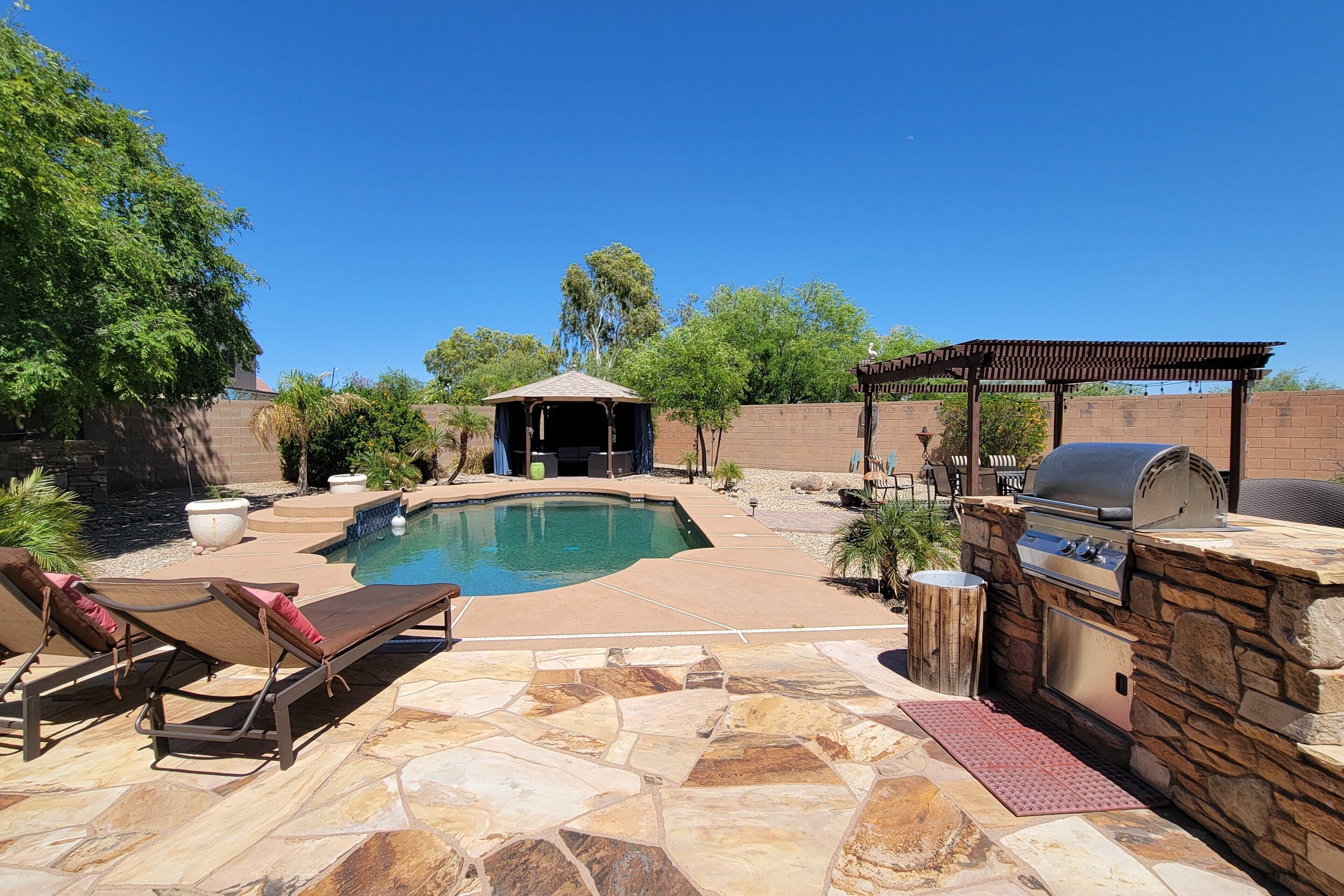 Un patio trasero con piscina, sillas de descanso, una parrilla y una pérgola.