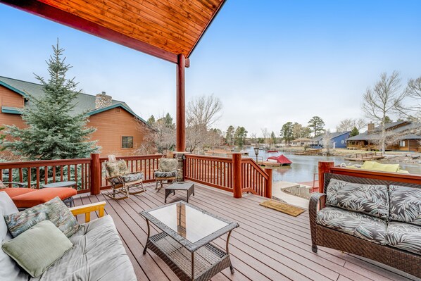 A wooden deck with outdoor furniture, a glass table, and a view of a lake and buildings in the background.