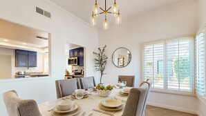 Comedor con una mesa blanca dispuesta para seis personas, rodeada de sillas grises tapizadas. Un candelabro cuelga encima, y se ve una ventana grande con persianas a la derecha.