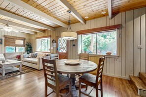 A wooden dining table with chairs, a ceiling fan, and a window with a view of trees.