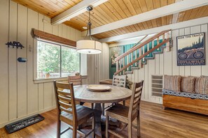 A wooden dining area with a round table, chairs, a hanging light fixture, a staircase, and a poster on the wall.