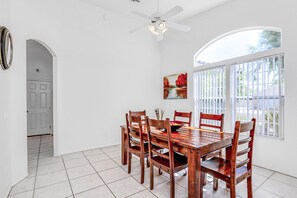 A dining area with a white tiled floor, a wooden table, and six chairs. There is a ceiling fan, a window with blinds, and a door leading to another room. - Peaceful, dog-friendly home with private pool, attached spa & patio (Palm Desert)