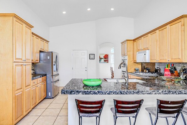 A kitchen with wooden cabinets, a stainless steel refrigerator, a granite countertop, a green bowl, and three bar stools. - Peaceful, dog-friendly home with private pool, attached spa & patio (Palm Desert)