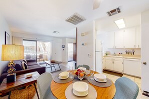 A modern living room with a sofa, dining table set for four, and a kitchen area with white cabinets and a sink. - Mountain view condo blocks from downtown with pool, tennis & pickleball (Palm Springs)