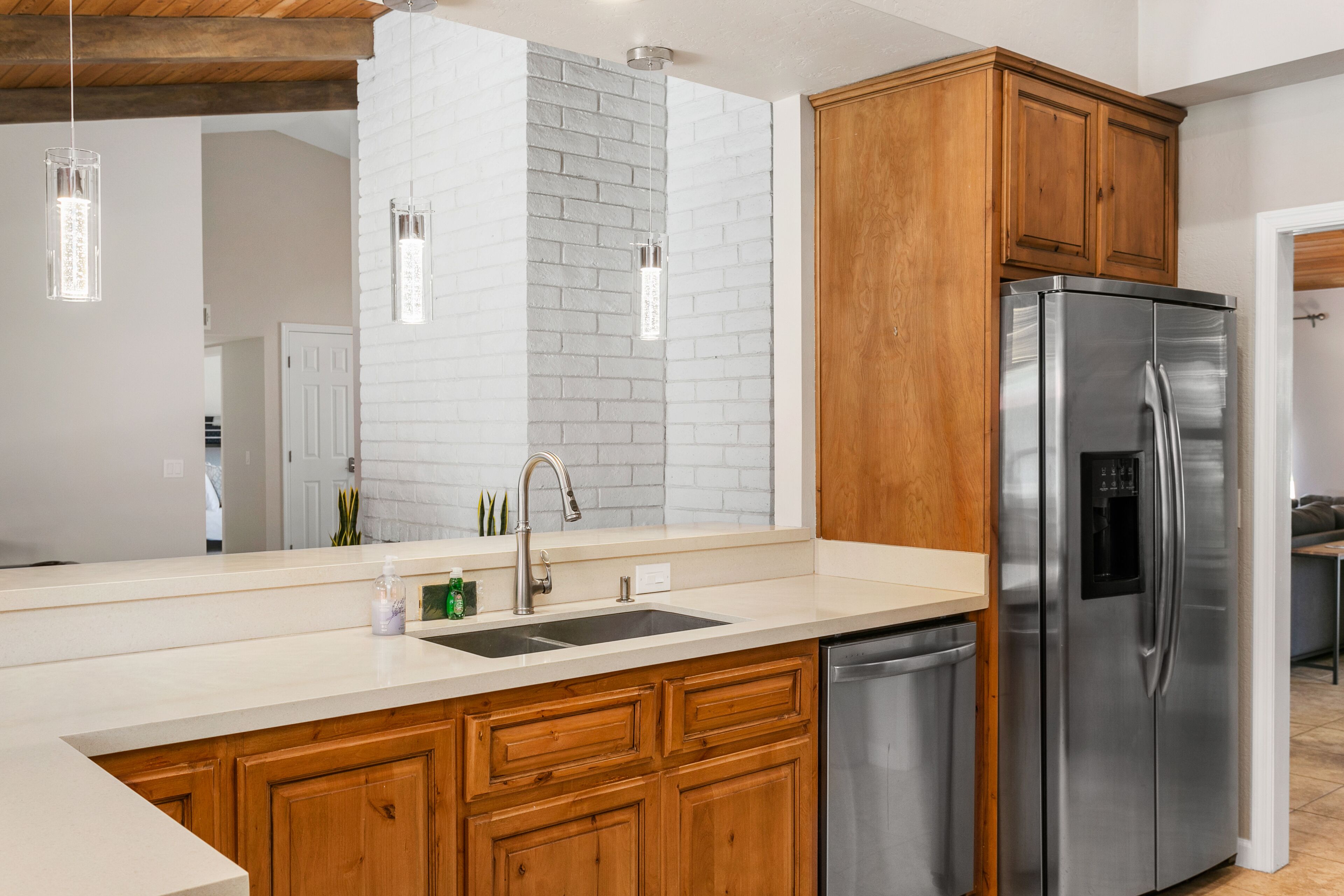 A modern kitchen with a stainless steel refrigerator, wooden cabinets, a white brick accent wall, and pendant lighting.