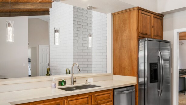 Una cocina moderna con refrigerador de acero inoxidable, armarios de madera, pared de ladrillo blanco y luminarias colgantes.