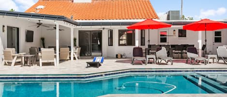 Una piscina con sillas de descanso y sombrillas, un área de patio cubierta y un edificio con tejado de tejas.