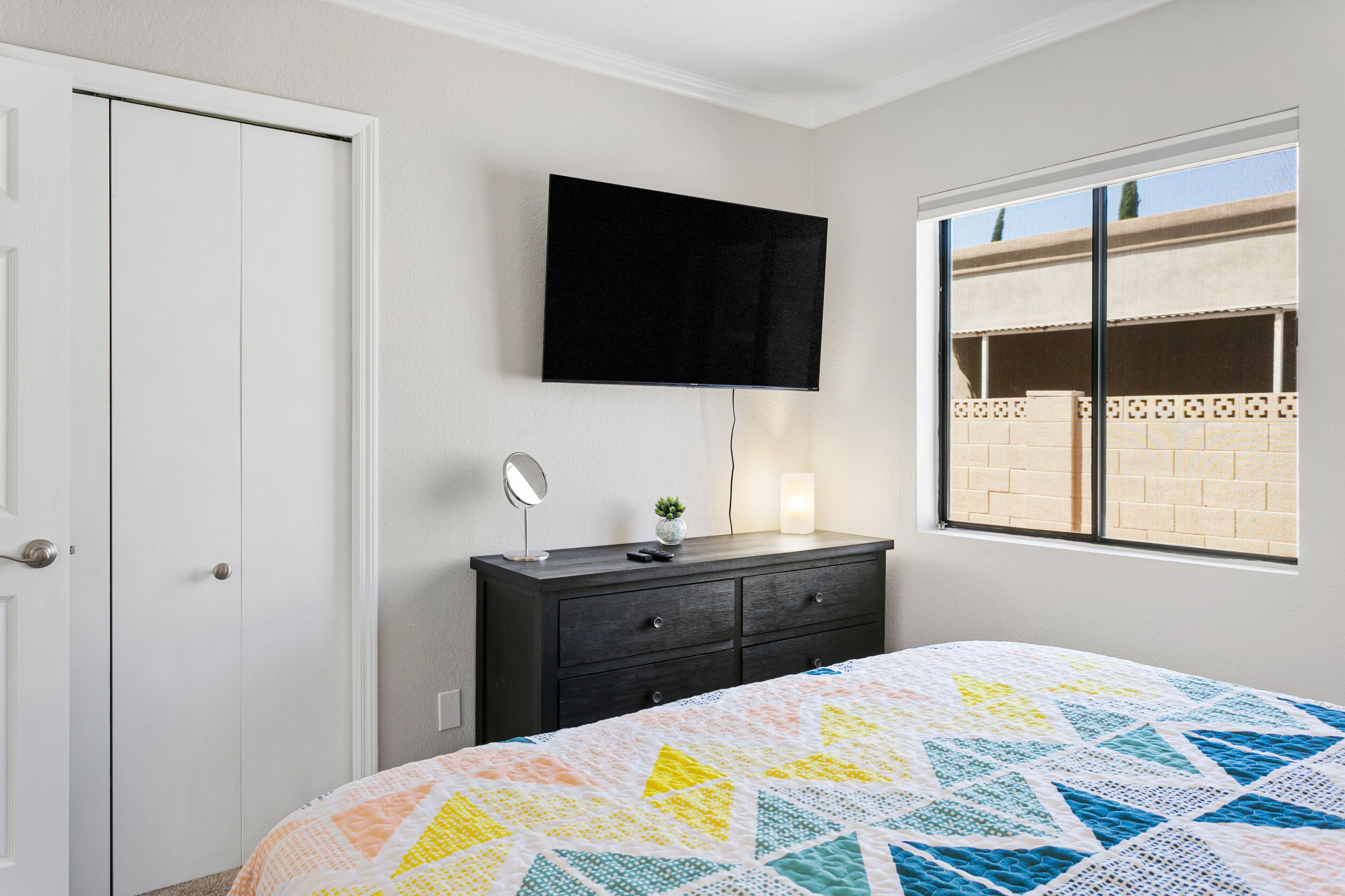 A bedroom with a bed, a dresser, a TV mounted on the wall, a window, and a door.