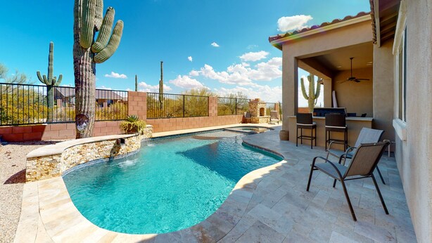A swimming pool with a curved edge, surrounded by a stone patio, and a covered seating area with chairs and a table.