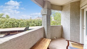 Un balcon avec un banc en bois, une voiture garée devant, et une vue sur la verdure et les bâtiments en arrière-plan.