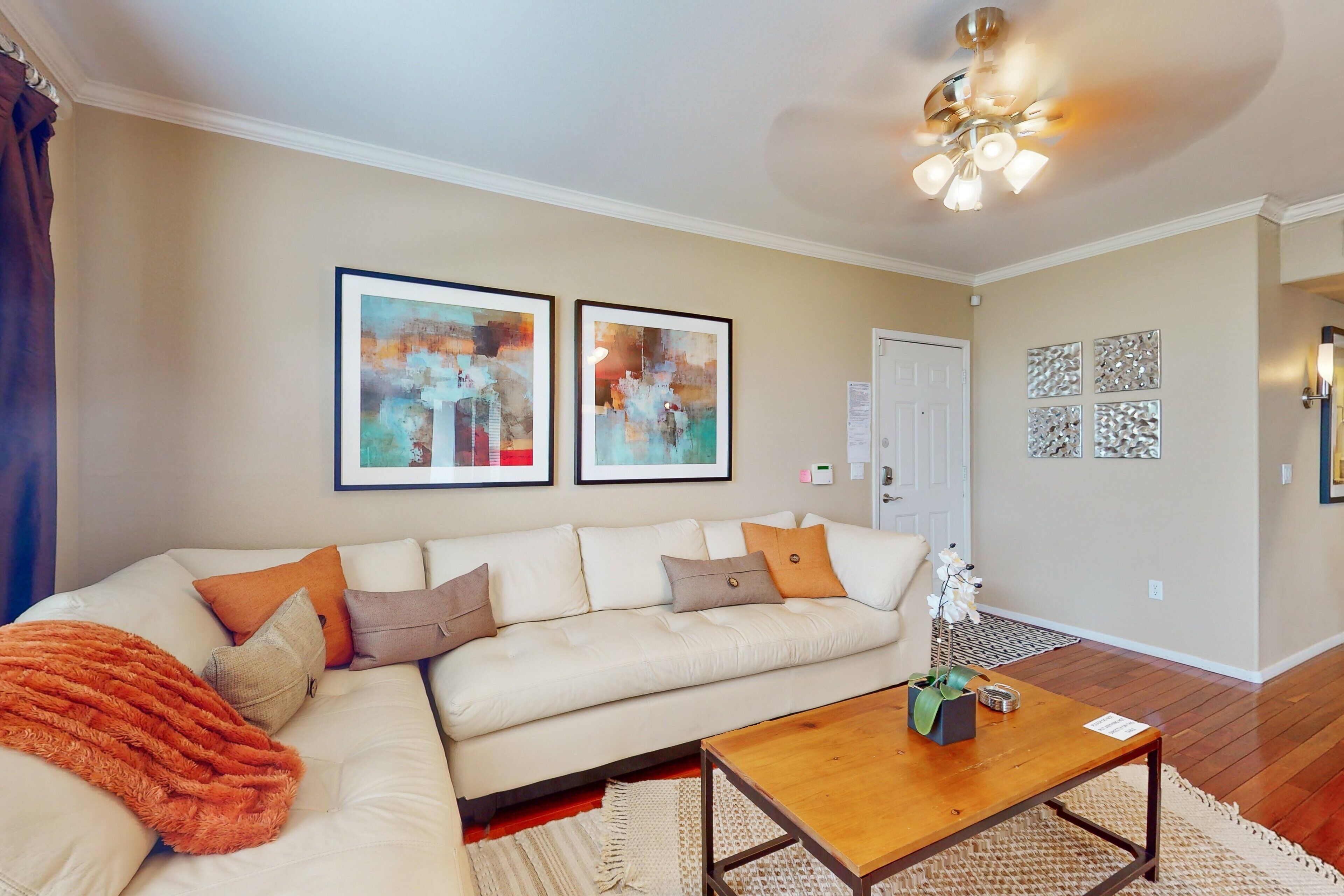 A living room with a beige sofa, a wooden coffee table, and a ceiling fan. There are abstract paintings on the wall and a door in the background.