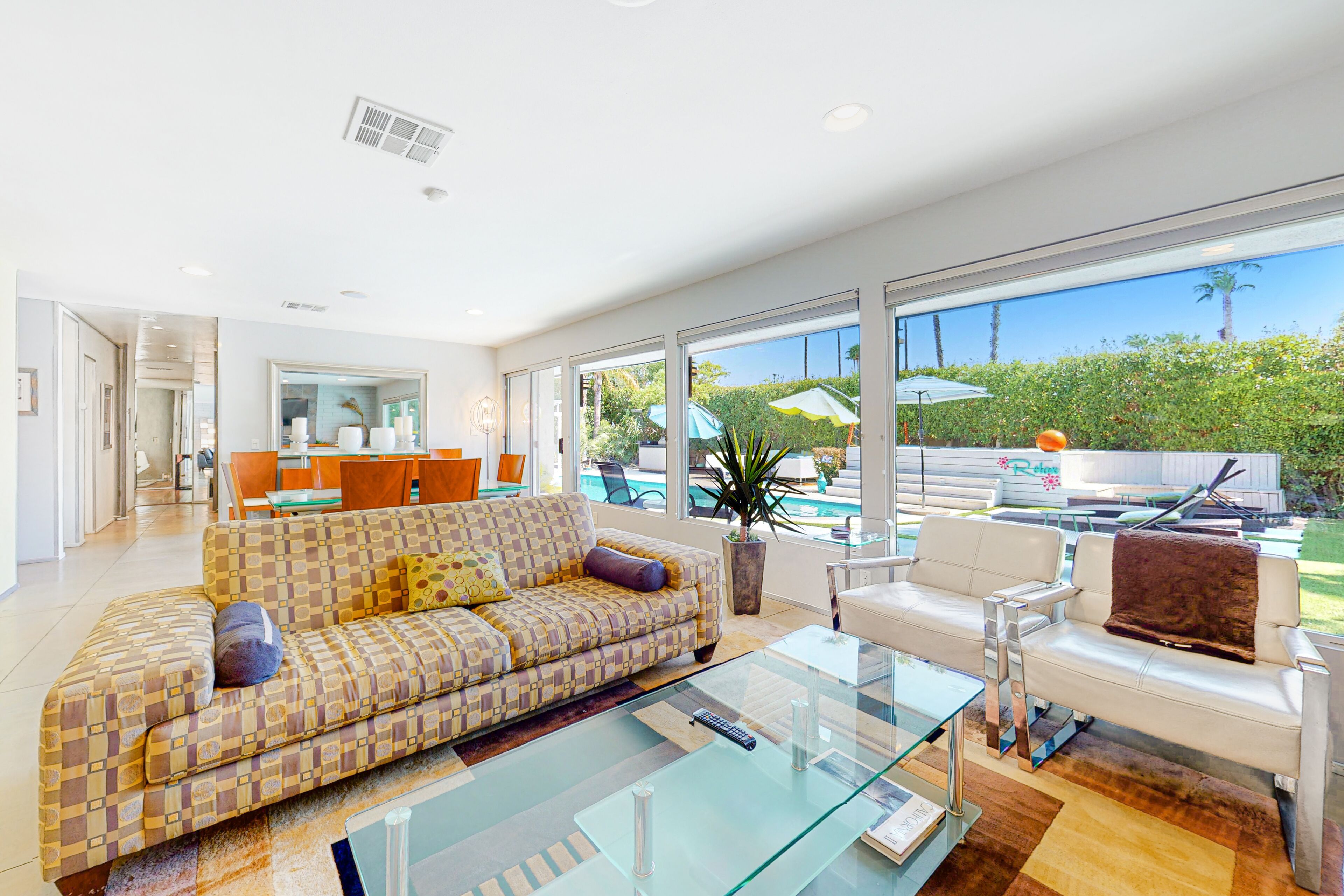 A spacious living room with a patterned sofa, a glass coffee table, and a white armchair. There is a large window offering a view of a pool and greenery.