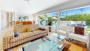 Ein großzügiges Wohnzimmer mit einer gemusterten Couch, einem Glastisch und einem weißen Sessel. Ein großes Fenster bietet Blick auf einen Pool und viel Grün.
