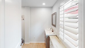 A narrow hallway with a white sink and mirror, a door on the left, and a window with blinds on the right. - Gorgeous Palm Desert Oasis with Backyard Paradise, Pool, Spa & Private Casita (Palm Desert)
