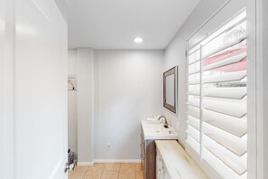 A narrow hallway with a white sink and mirror, a door on the left, and a window with blinds on the right. - Gorgeous Palm Desert Oasis with Backyard Paradise, Pool, Spa & Private Casita (Palm Desert)