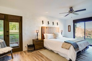 A bedroom with a large bed, a wooden nightstand, a ceiling fan, and a view of the outdoors through a sliding door. - Mountain-view retreat with amazing views of Palm Desert city lights & pool (Palm Desert)