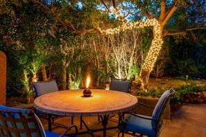A round outdoor table with chairs, surrounded by greenery and illuminated by string lights. - Mountain-view retreat with amazing views of Palm Desert city lights & pool (Palm Desert)