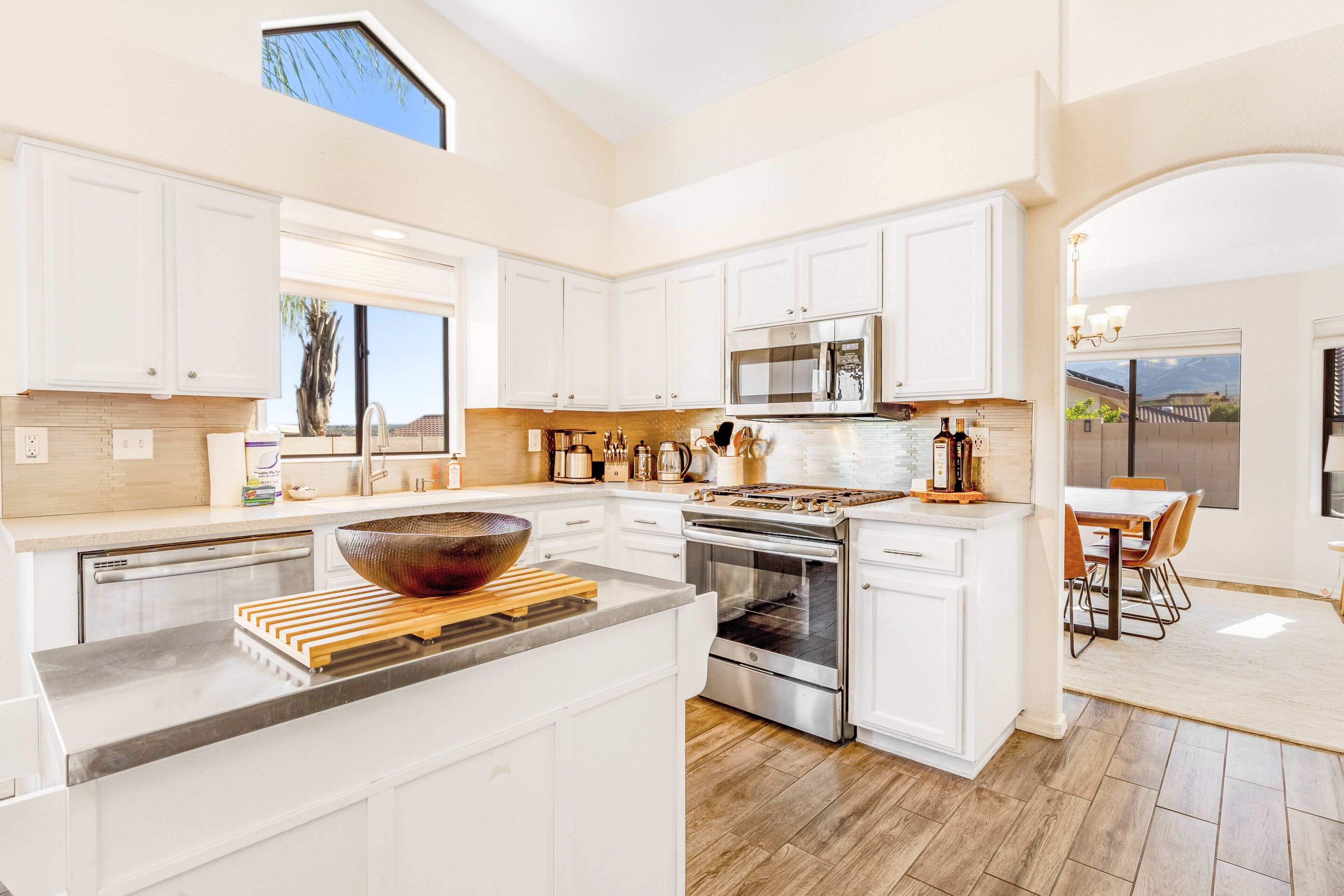 A modern kitchen with white cabinets, a large island, stainless steel appliances, and a dining area with a view.