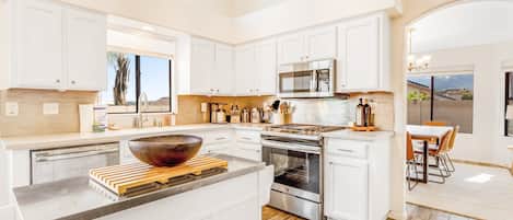 A modern kitchen with white cabinets, a large island, stainless steel appliances, and a dining area with a view.
