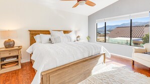 Een slaapkamer met een houten bed, een plafondventilator, een groot raam met uitzicht op bergen, en een witte bank.