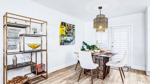 Un espace repas doté d’un plancher en bois, de chaises blanches, d’une table à plateau en verre et d’une étagère en métal décorative où sont exposés divers objets.