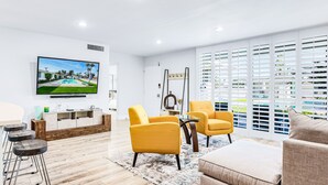 Un salon moderne avec un téléviseur, des fauteuils jaunes, un canapé et une grande fenêtre équipée de stores.
