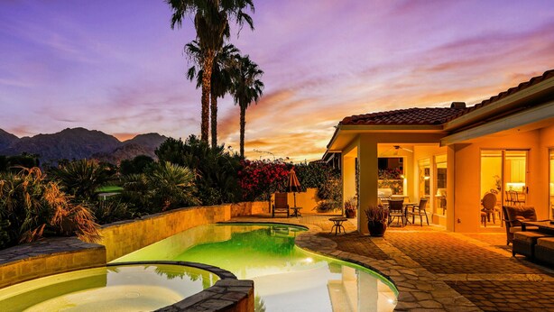 Ein Schwimmbecken mit Steinrand, umgeben von einer Terrasse mit Gartenmöbeln und Blick auf die Berge bei Sonnenuntergang.