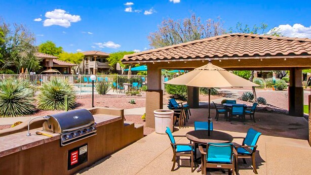 En udendørs terrasse med grill, siddepladser og en pool i baggrunden.
