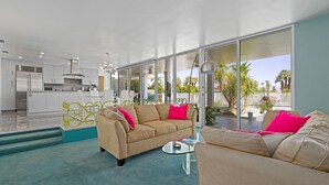 Una sala moderna con un sofá color beige, una mesa de centro de vidrio y una cocina al fondo.