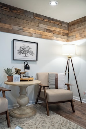 Ein Eckzimmer mit Holzbalkendecke, einem gerahmten Baum-Kunstwerk, einer Stehlampe, einem runden Tisch mit Topfpflanze, einem Sessel und einem kleinen Teppich.