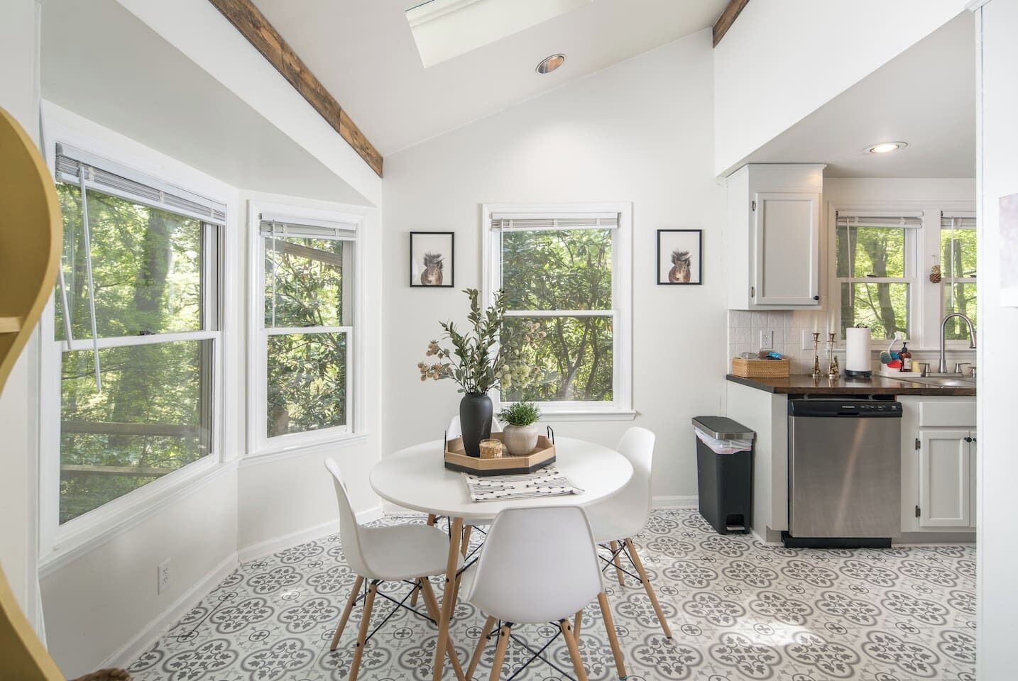 Une cuisine moderne avec des meubles blancs, un coin repas doté d’une table ronde et de chaises, et de grandes fenêtres offrant une vue sur les arbres.