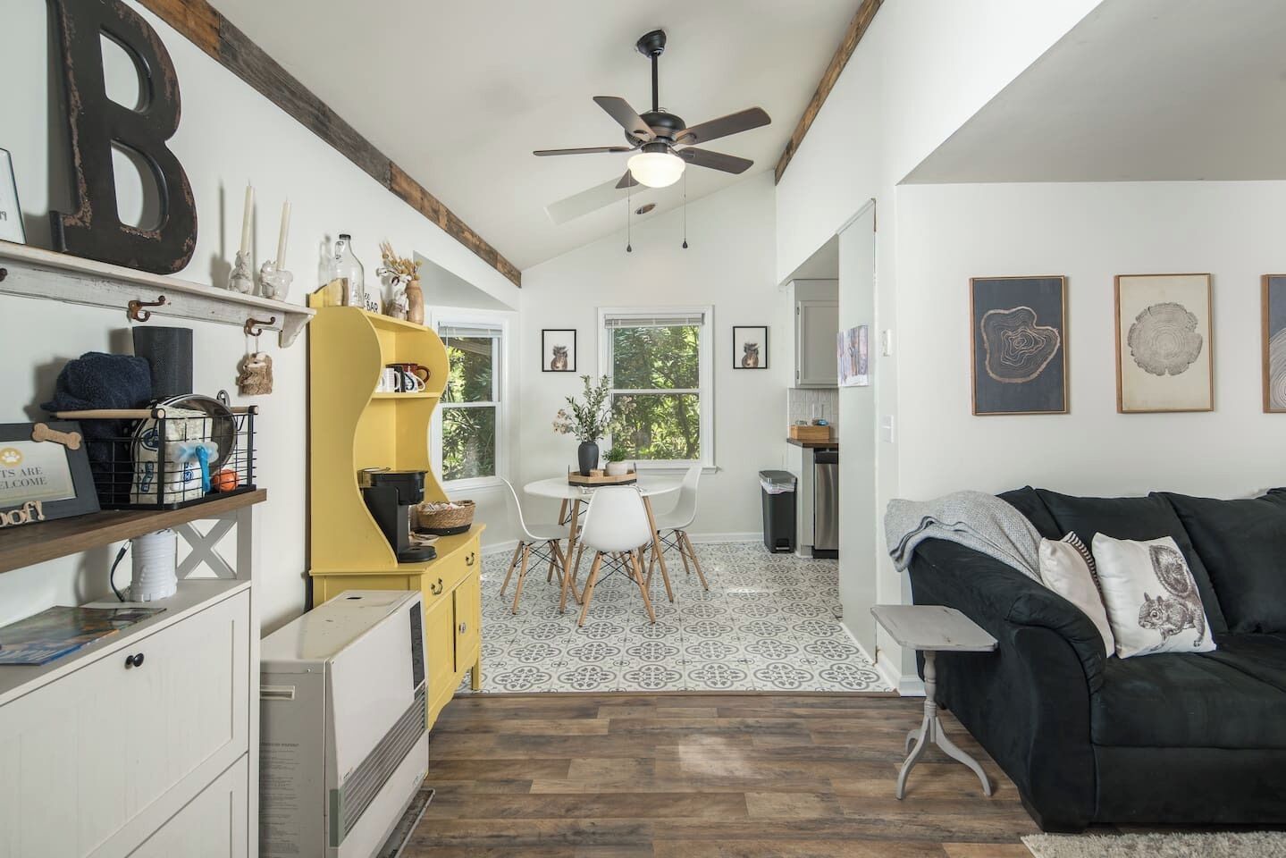 Een moderne keuken met een gele accentmuur, een eethoek met een witte tafel en stoelen, en een zwarte bank met decoratieve kussens.