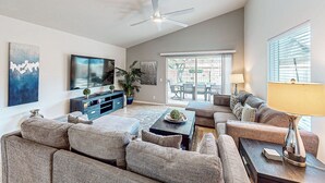 Uma sala de estar espaçosa com um amplo sofá em formato de L, uma mesa de centro, uma TV e um ventilador de teto.