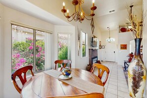 A dining area with a wooden table, chairs, a vase with dried branches, and a view of pink flowers through the window. - Dog-Friendly Oro Valley Retreat | Private Hot Tub, Catalina Views & Shaded Patio (Oro Valley)