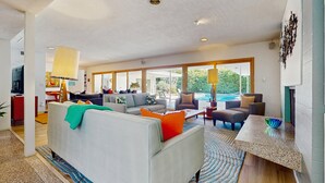 Un salón amplio con un sofá grande, varios sillones y una mesa de centro. Se ve una piscina a través de amplias ventanas.