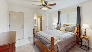 A bedroom with a wooden bed, a ceiling fan, a window with curtains, and a bedside table with a lamp.