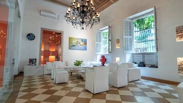 A spacious living room with a checkered floor, white furniture, a chandelier, and a large window with shutters.