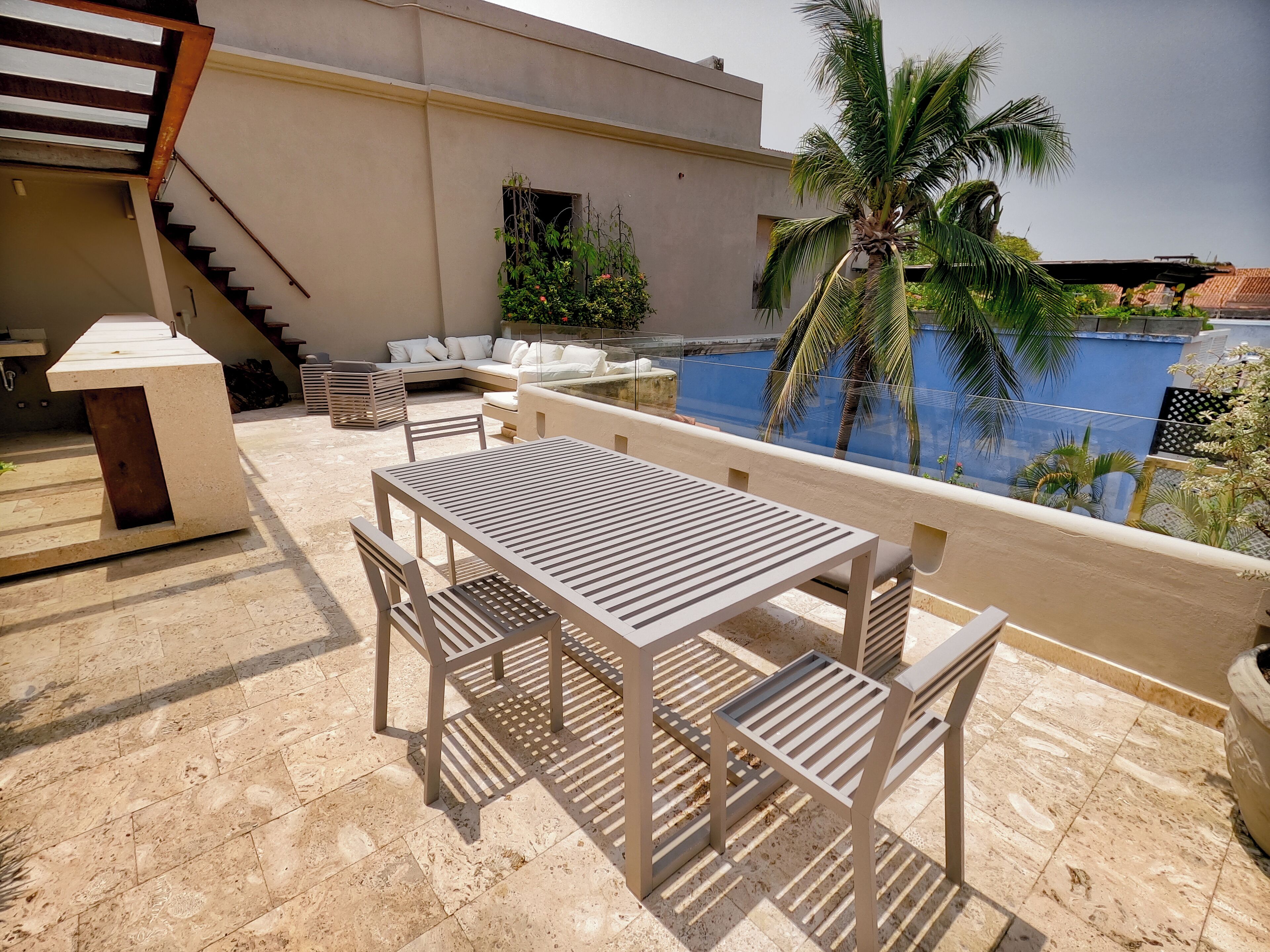 A terrace with a table and chairs, a staircase, and a view of a blue building and palm trees.