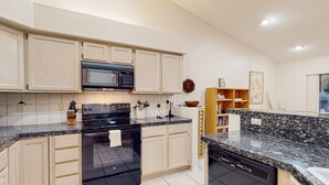 Una cocina moderna con armarios de madera clara, microondas integrado, horno y lavavajillas, encimeras de granito y una pequeña isla con fregadero.
