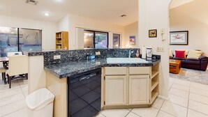 Cocina moderna con encimeras de granito, lavavajillas y armarios de madera. Cuenta con un comedor con mesa y sillas, y una sala de estar con sofá y mesa de centro.