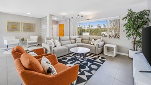 A modern living room with a sectional sofa, orange armchairs, a coffee table, and a large window with a view of palm trees. - Mid-century modern home with private pool, spa, grill & mountain views (Palm Springs)