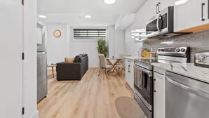 Una cocina moderna con electrodomésticos de acero inoxidable, una zona de comedor con mesa y sillas, y una sala de estar con sofá y una planta.