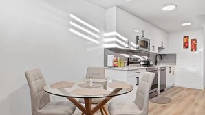 Una cocina moderna con armarios blancos, un microondas y una estufa. En primer plano, hay una mesa de vidrio redonda con cuatro sillas.