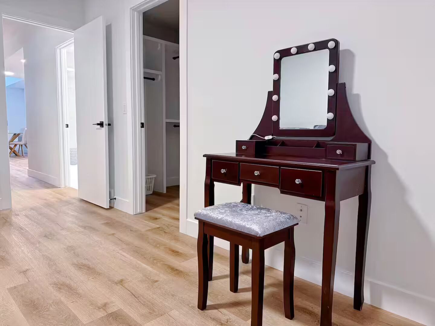 Un tocador de madera con espejo e iluminación, un banco y una puerta que da a un pasillo.