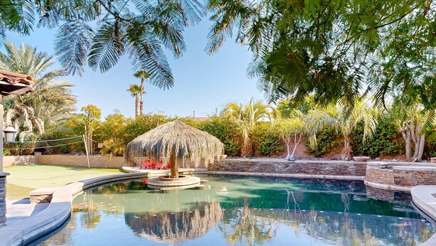 A swimming pool with a thatched-roof structure, surrounded by palm trees and a stone wall. - Beautiful home with private pool, outdoor kitchen & enclosed yard (Indio)
